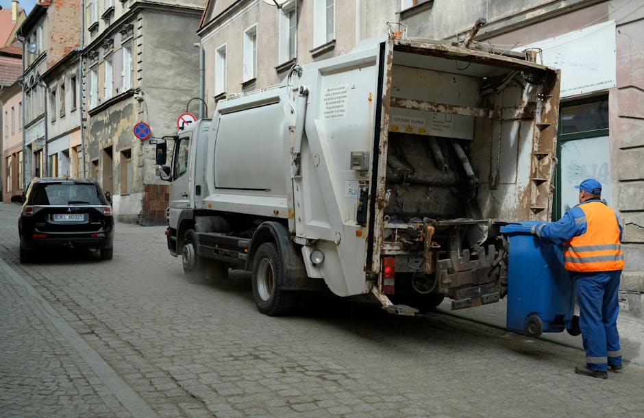 A large open-top recycling and waste bin station situated outside a commercial building on a paved street, with various types of rubbish overflowing onto the surrounding pavement. The central grey bin