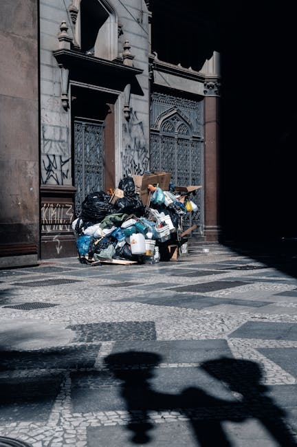 A photograph of a large, open flatbed truck loaded with various types of waste materials parked on a paved surface in an outdoor area. The waste includes several cardboard boxes, some visibly crushed 